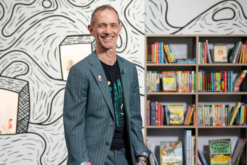 Andy Griffiths stands in front of an exhibition of objects displayed through windows in a swirl-patterned background.