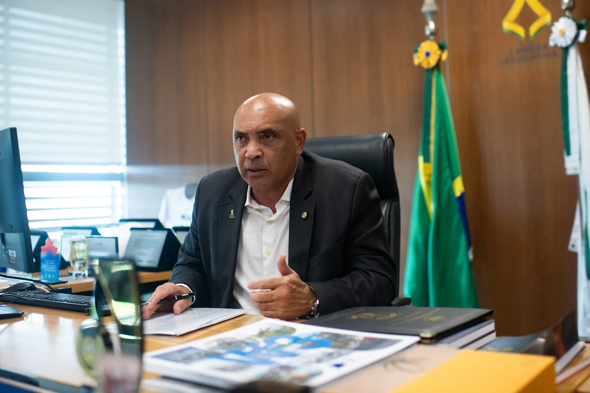 Foto de homem sentado em cadeira com mesa à frente e bandeira ao fundo